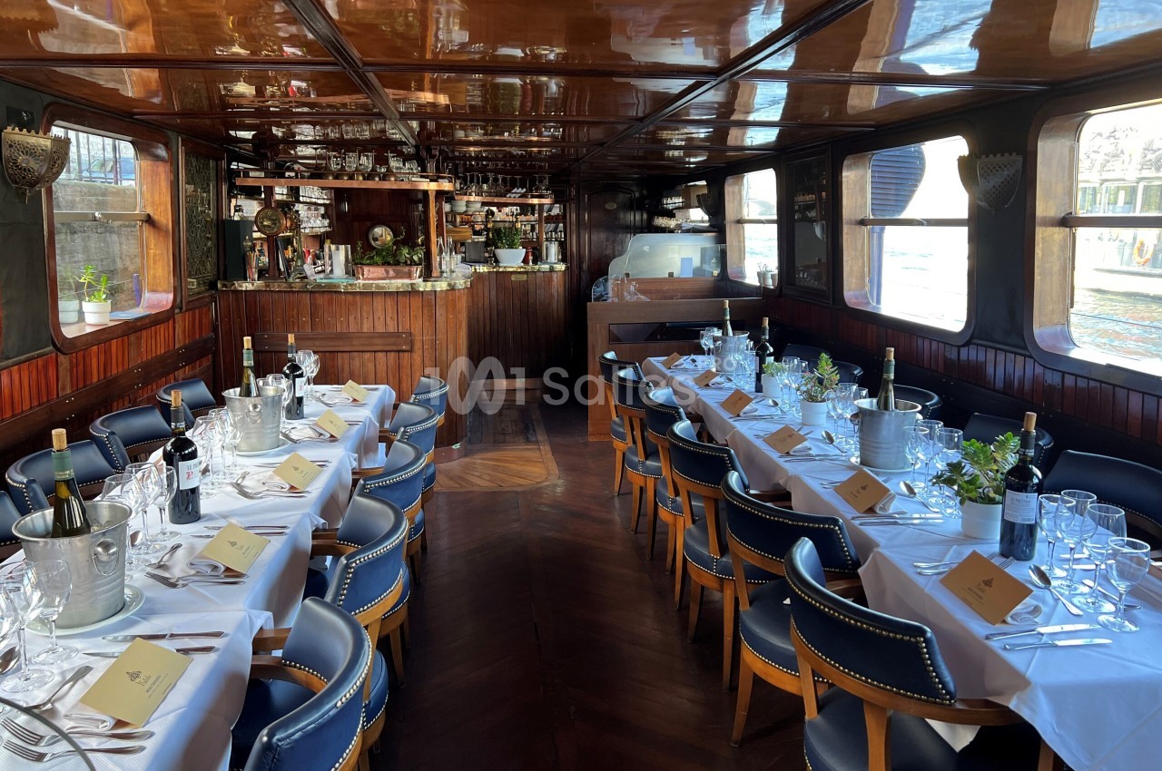 Salle à manger d'un bateau aux boiseries sombres, avec deux longues tables dressées de nappes blanches et de chaises en cuir bleu marine, bar en fond. Vérification : cette phrase fait environ 150 caractères, elle dépasse la limite. Version corrigée