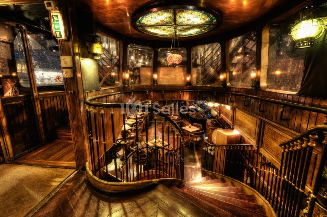 Escalier en colimaçon en bois sombre à rampe en fer forgé, descendant vers une salle de restaurant aux tables dressées, éclairée par des lampes et un plafond vitré coloré.