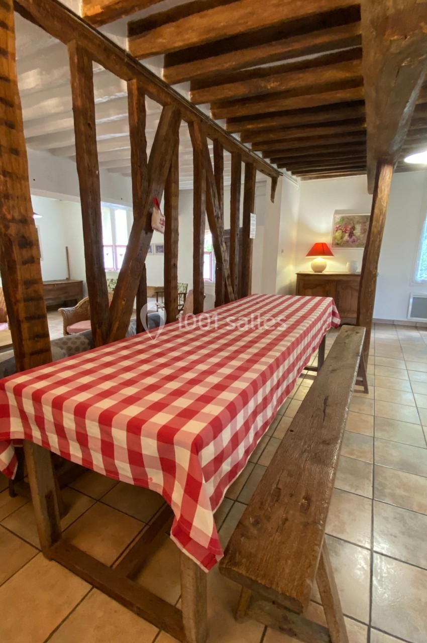 Table en bois avec nappe à carreaux rouges et blancs, entourée de bancs, dans une pièce rustique avec poutres apparentes.
