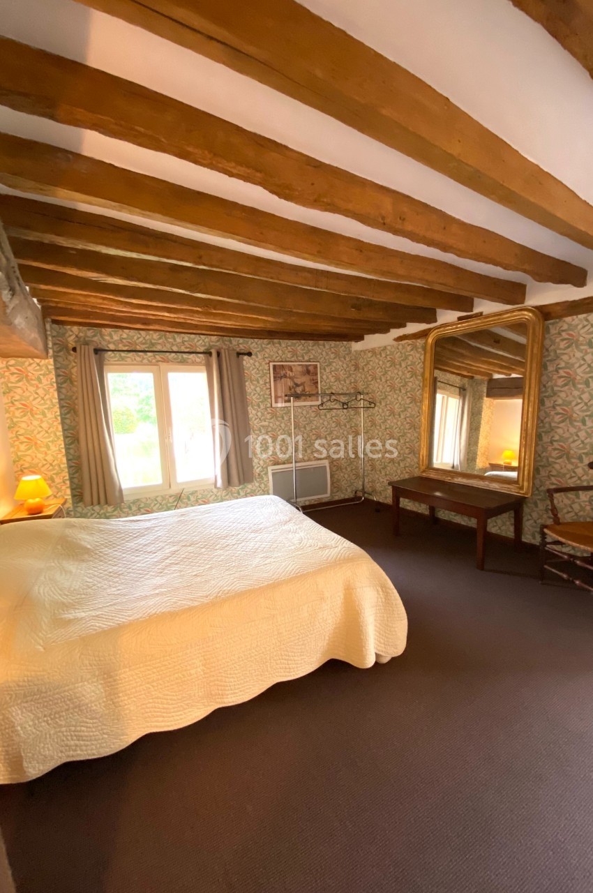 Chambre lumineuse avec poutres apparentes, lit double, miroir doré, chaise en bois et vue sur une fenêtre.