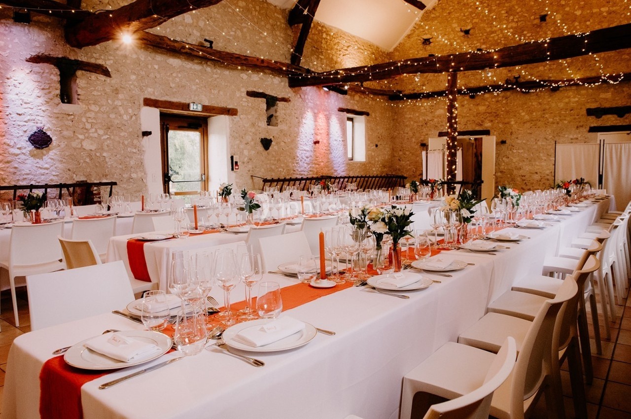 Salle de réception décorée avec des tables alignées, nappes blanches, chemins de table rouges et éclairage chaleureux.