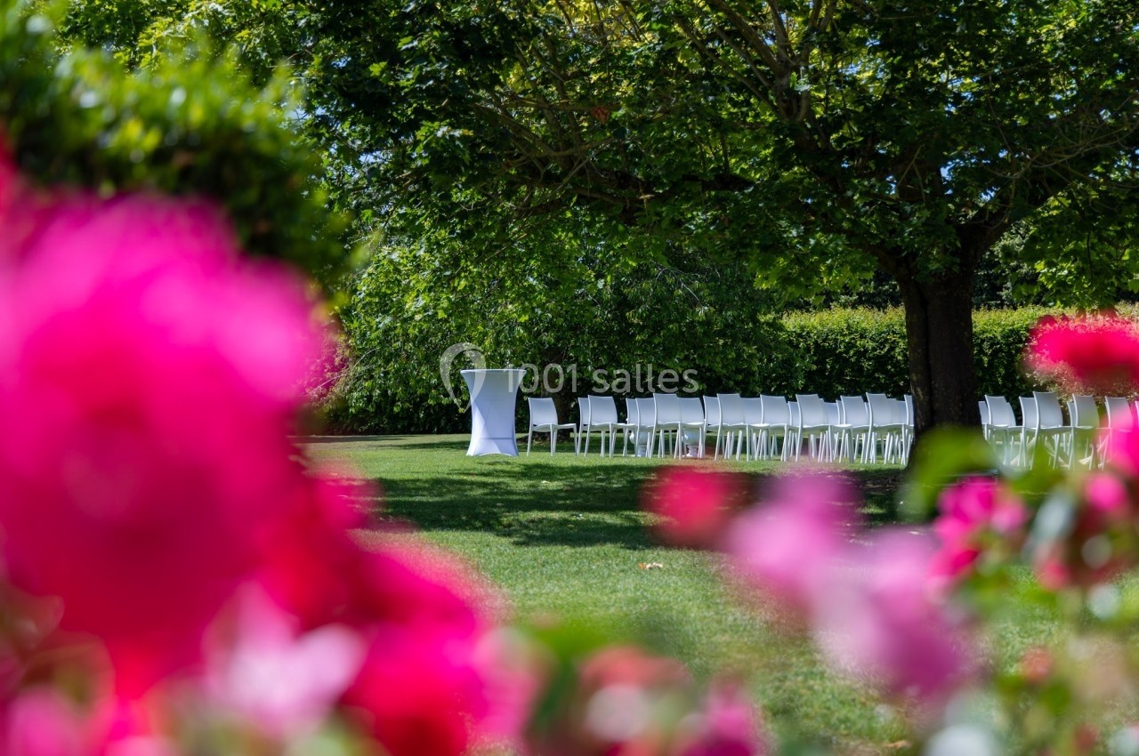 Chaises blanches alignées sous un grand arbre dans un jardin, avec des fleurs roses floues au premier plan.