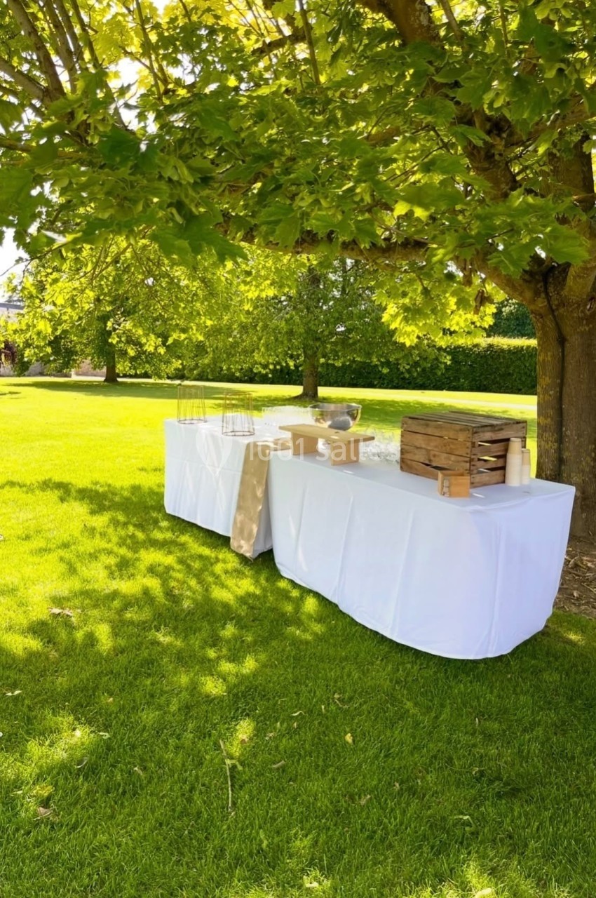 Table dressée sous un arbre dans un jardin, avec nappes blanches et éléments de service en bois et métal.