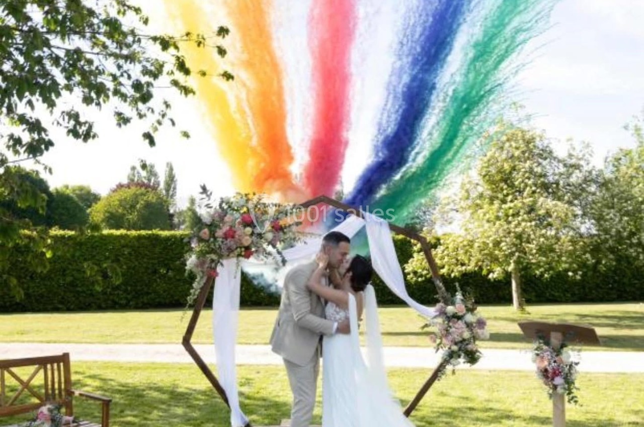 Un couple s'embrasse sous une arche décorée, avec des jets de poudre colorée en arrière-plan dans un jardin.