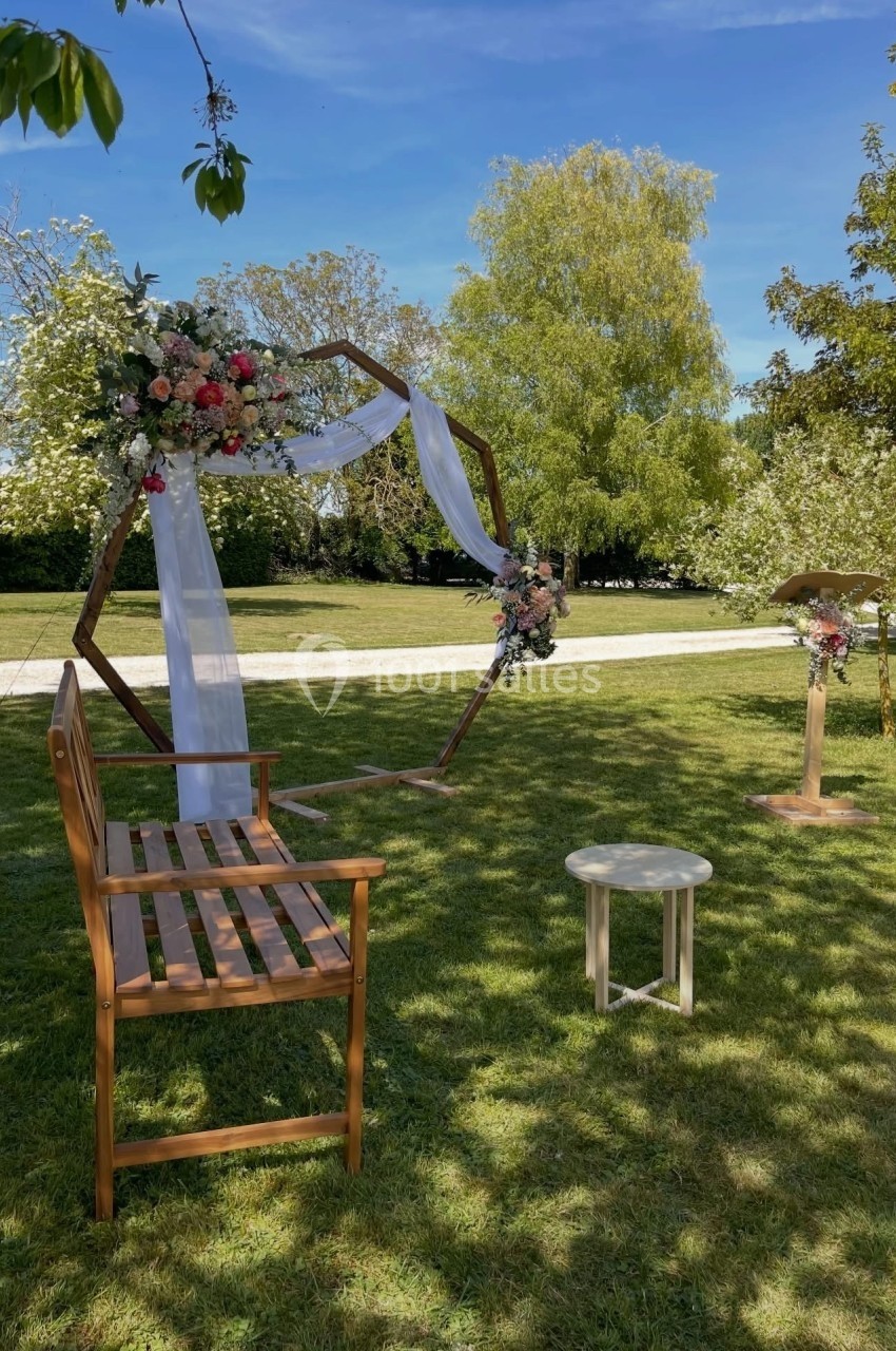 Arche hexagonale décorée de fleurs et de voilages blancs dans un jardin, avec banc en bois et pupitre sur l'herbe.