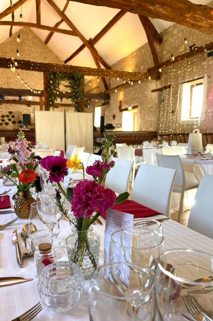 Salle de réception décorée pour un événement, avec tables dressées, fleurs colorées et guirlandes lumineuses suspendues.