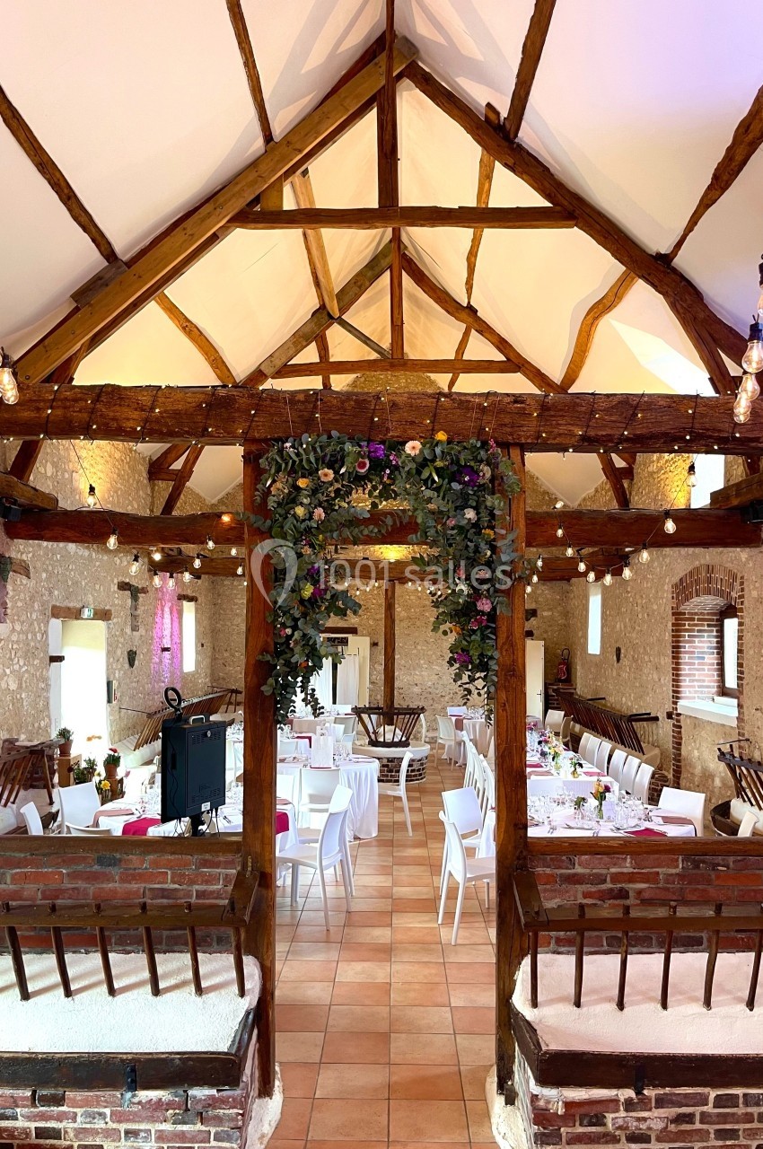 Salle de réception rustique avec poutres apparentes, tables dressées et décoration florale suspendue.