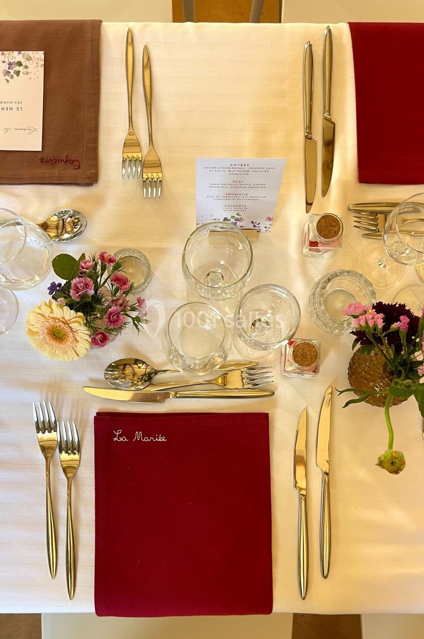 Table dressée avec nappes blanches, serviettes rouges et brunes, couverts dorés, verres et décorations florales.