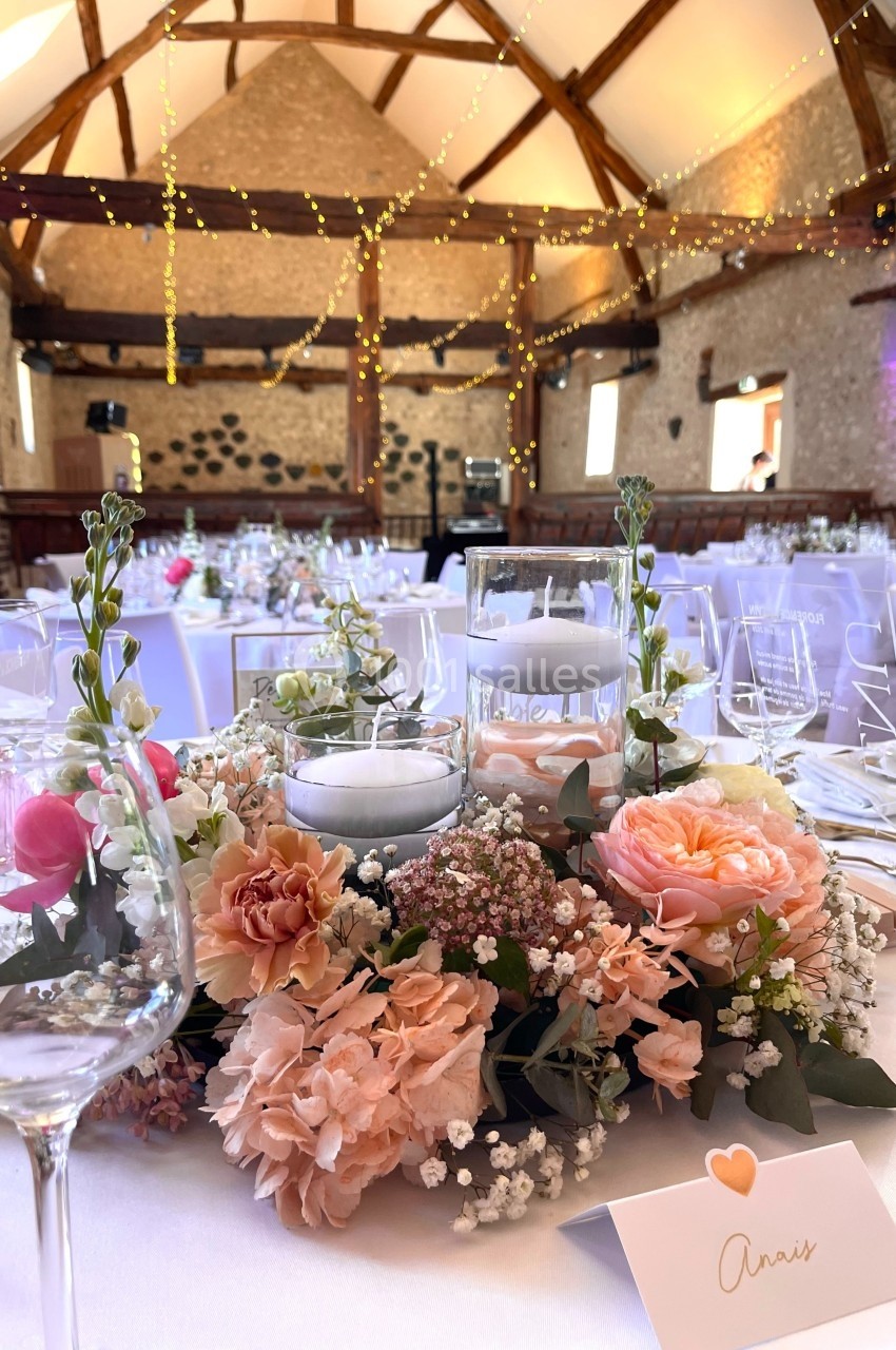 Centre de table floral avec bougies, entouré de verres et nappes blanches, dans une salle en pierres décorée de guirlandes…