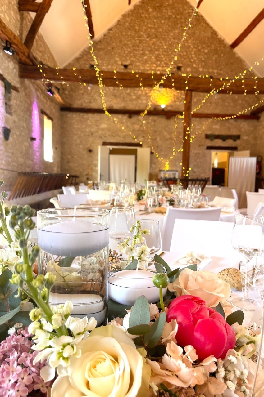 Salle de réception décorée pour un événement, avec tables dressées, fleurs et guirlandes lumineuses suspendues.