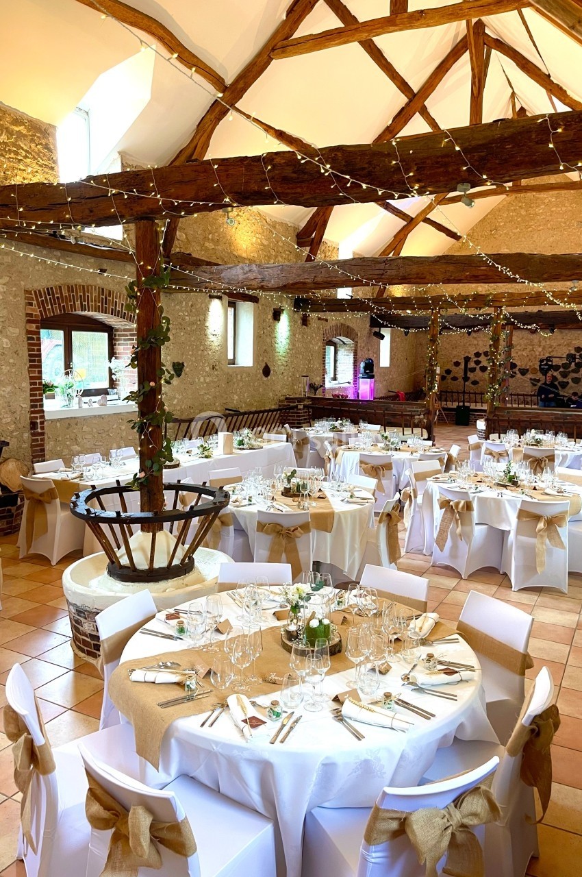 Salle de réception décorée pour un mariage, avec tables rondes dressées, nappes blanches et guirlandes lumineuses suspendues.