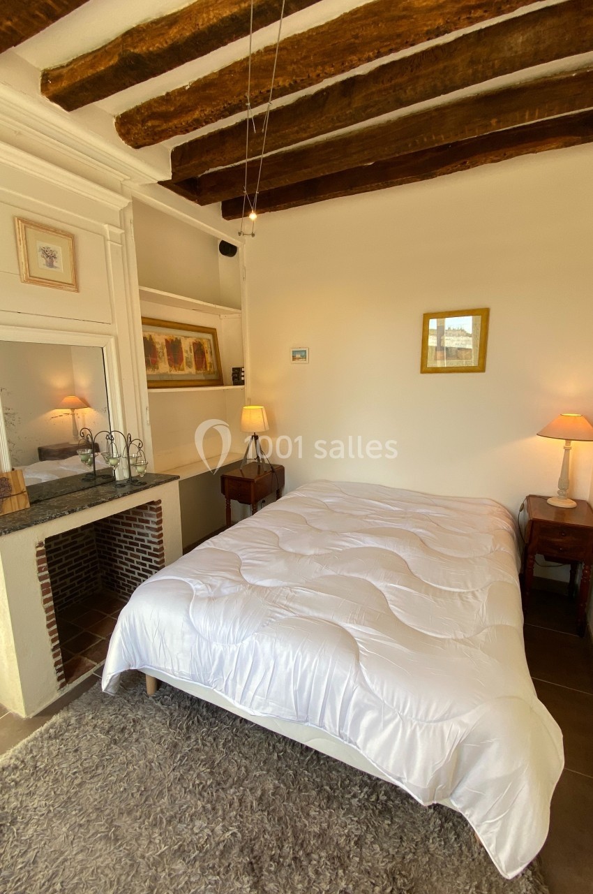 Chambre lumineuse avec poutres apparentes, lit double, tables de chevet, lampes et cheminée décorative.