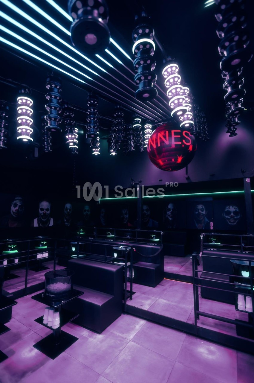 Salle de club aux tons violets et bleus, avec banquettes noires, tables basses, néons en lignes au plafond et grande sphère lumineuse rouge suspendue.
