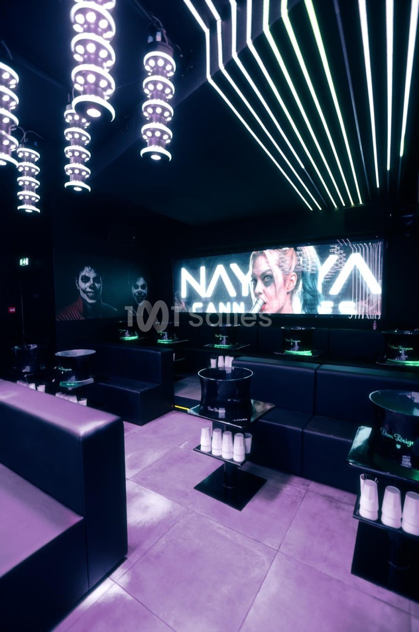 Intérieur de club sombre aux banquettes noires et tables hautes éclairées par des néons verts, avec un grand écran lumineux au mur et des luminaires LED suspendus.