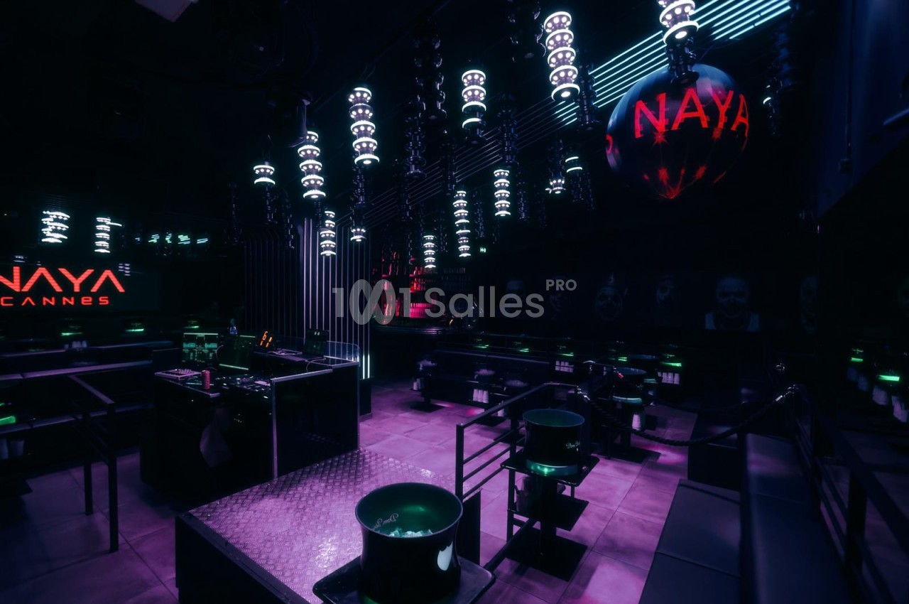 Club à l'ambiance sombre éclairée de violet, avec lampes suspendues, banquettes et seaux à glace sur tables basses.