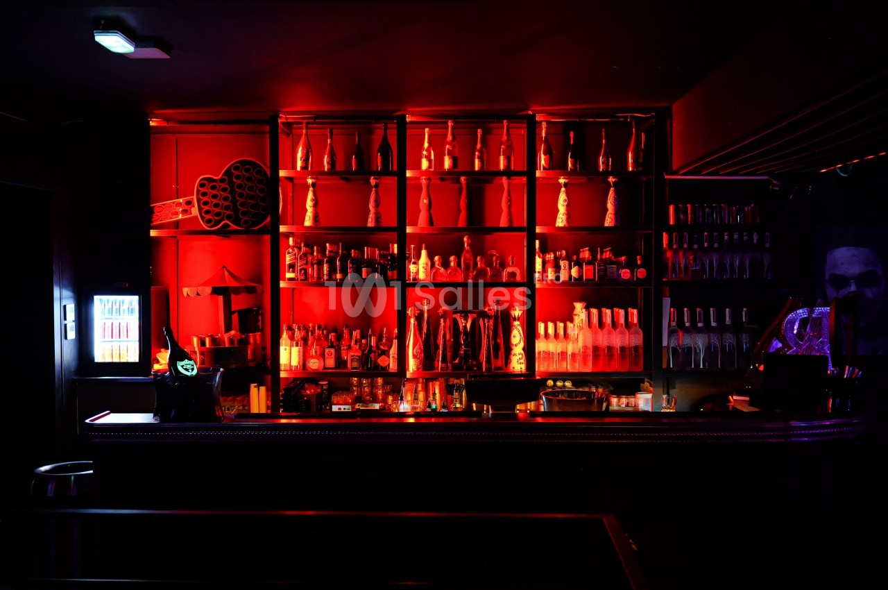 Bar en métal aux étagères éclairées en rouge, garnies de bouteilles d'alcool et de verres, dans une salle à l'ambiance sombre.