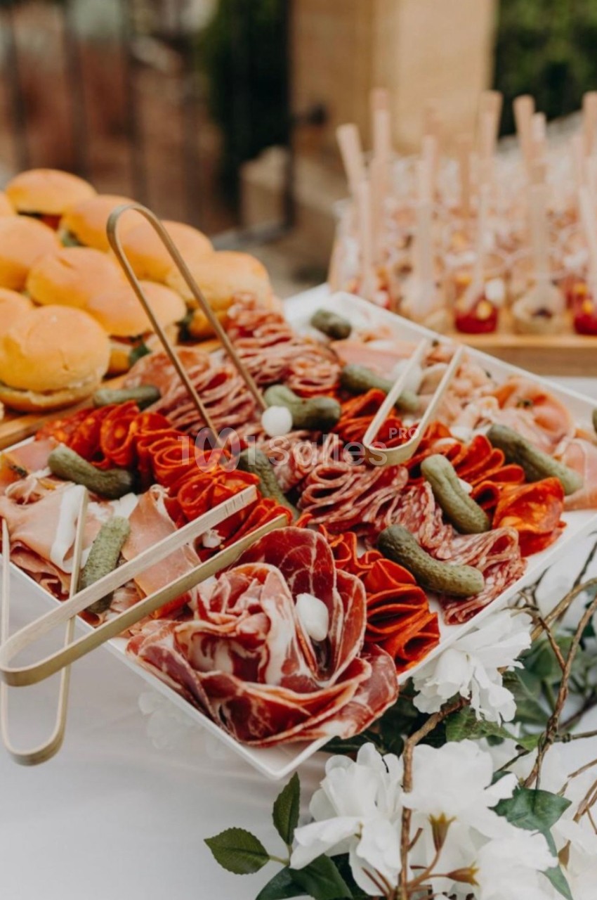 Plateau de charcuteries en rosettes (saucisson, jambon cru, bresaola) accompagné de cornichons, sur une table dressée en extérieur, avec des petits pains et des verrines à l'arrière-plan.