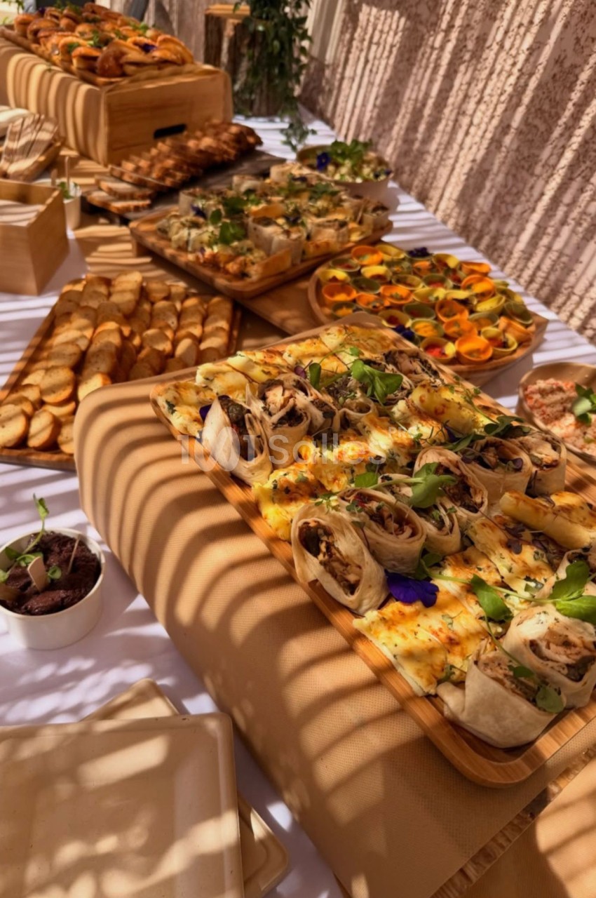 Buffet de pièces salées sur planches en bois : wraps gratinés garnis de viande, toasts, verrines colorées décorées de fleurs comestibles, sous la lumière du jour.