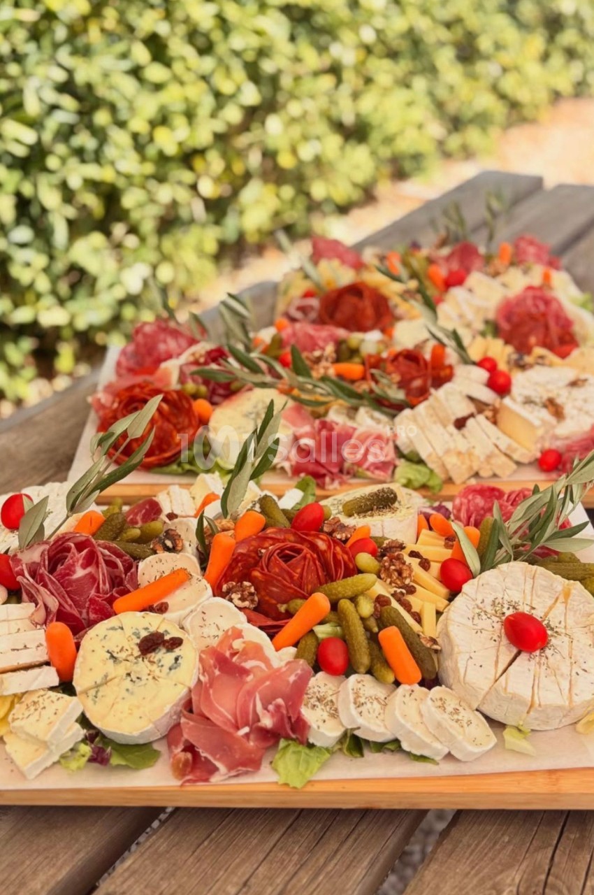 Plateaux de charcuteries, fromages, cornichons, carottes et tomates cerises disposés sur des planches en bois, en extérieur devant une haie verte. Count characters: roughly 135? Let's count roughly: "Plateaux de charcuteries, fromages, cornichons