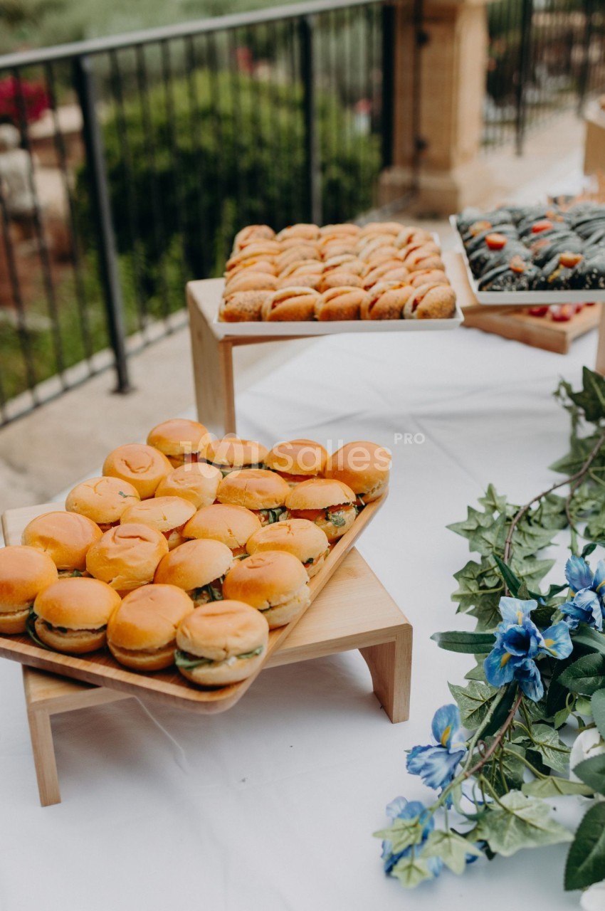 Plateaux de mini-sandwichs sur pain rond et viennoiseries en pâtisserie disposés sur une table nappée de blanc, en extérieur près d'une balustrade, avec une guirlande de fleurs bleues. Let me check length: roughly 180 characters, too long. Need ≤125.
