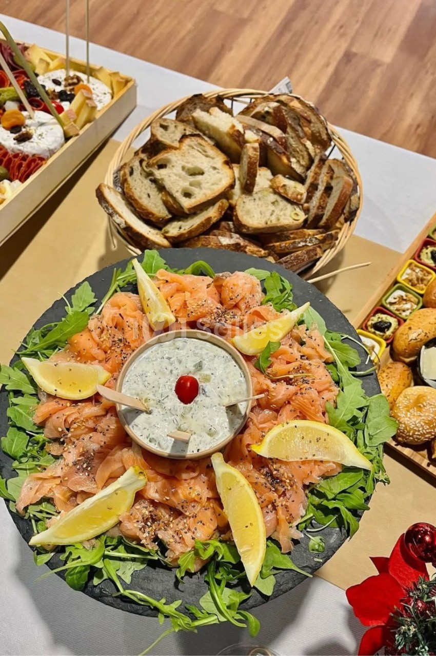 Plateau rond en ardoise de saumon fumé fin disposé sur roquette, garni de quartiers de citron et d'une coupelle de sauce crémeuse aux herbes, à côté d'un panier de pain et de canapés. Note : la phrase fait environ 190 caractères, au-delà de la limite.