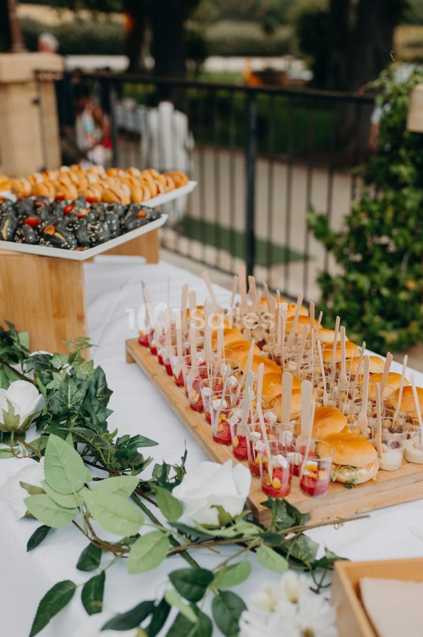 Plateau en bois de mini-sandwiches et de verrines garnies de maïs sur une table nappée de blanc, en extérieur.