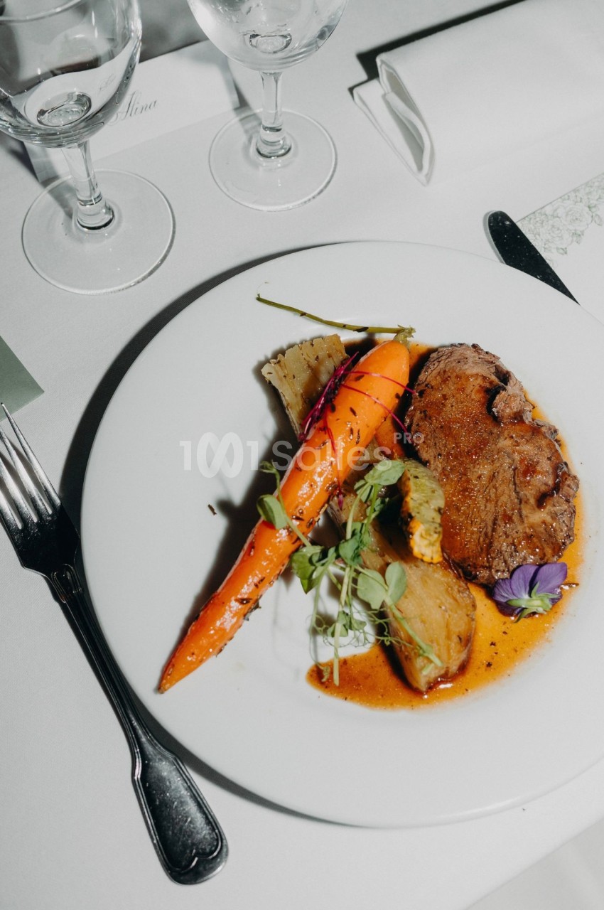 Assiette dressée sur nappe blanche : pièce de viande rôtie nappée de jus, carotte glacée, légumes braisés, pousses vertes et fleur violette, couverts et verres à pied à proximité. Vérification : environ 150 caractères, trop long. Reformulation : Assiette