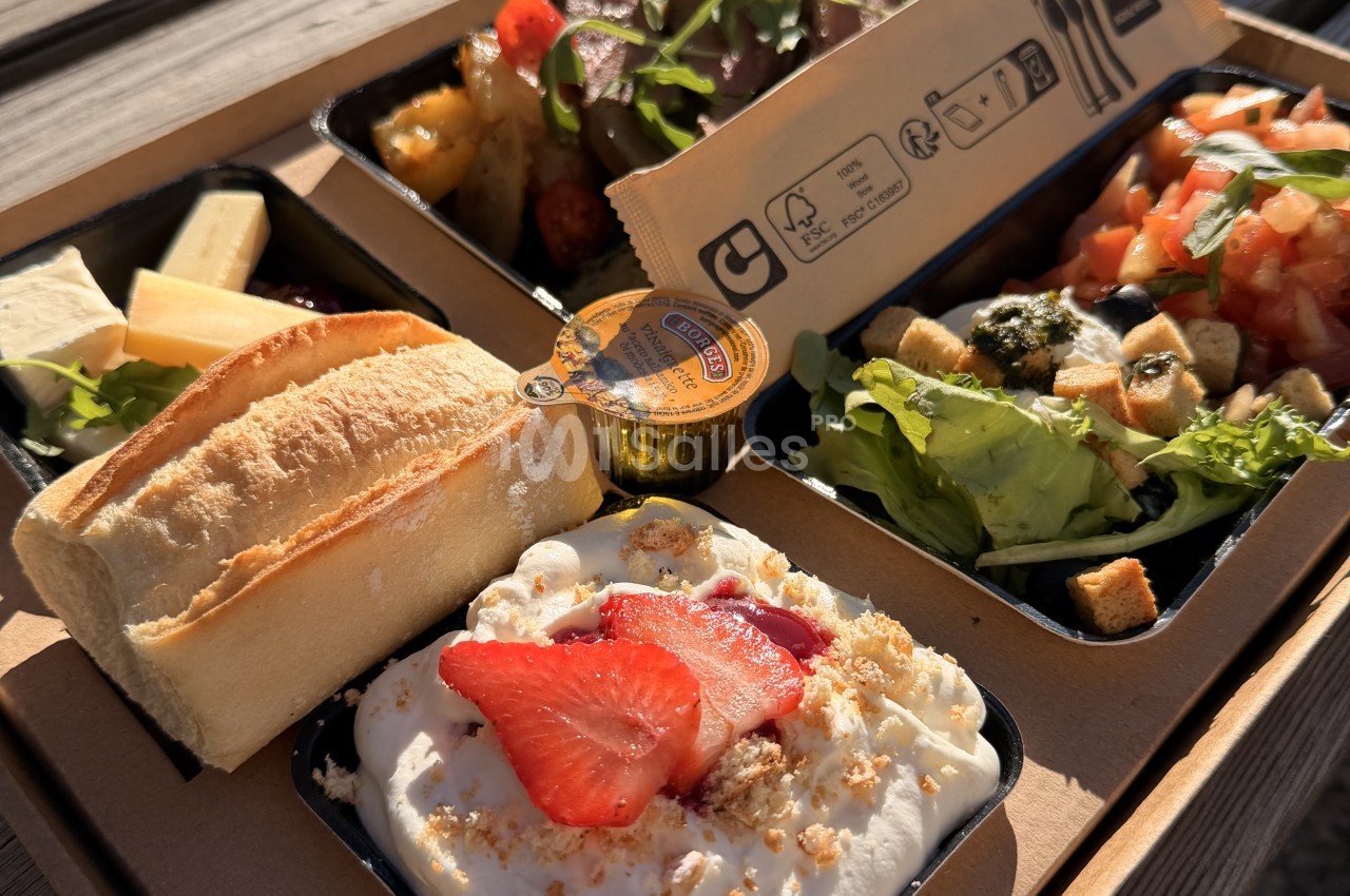 Panier-repas en carton ouvert présentant un baguette, des fromages, une salade de tomates et croûtons, une salade verte, une salade de pommes de terre et un dessert crémeux garni de fraises.