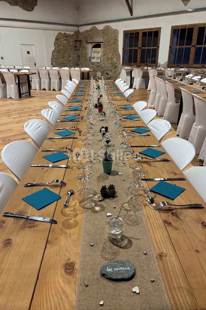 Grande table en bois décorée pour un repas, avec chemins de table, bougies, serviettes bleues et chaises blanches.