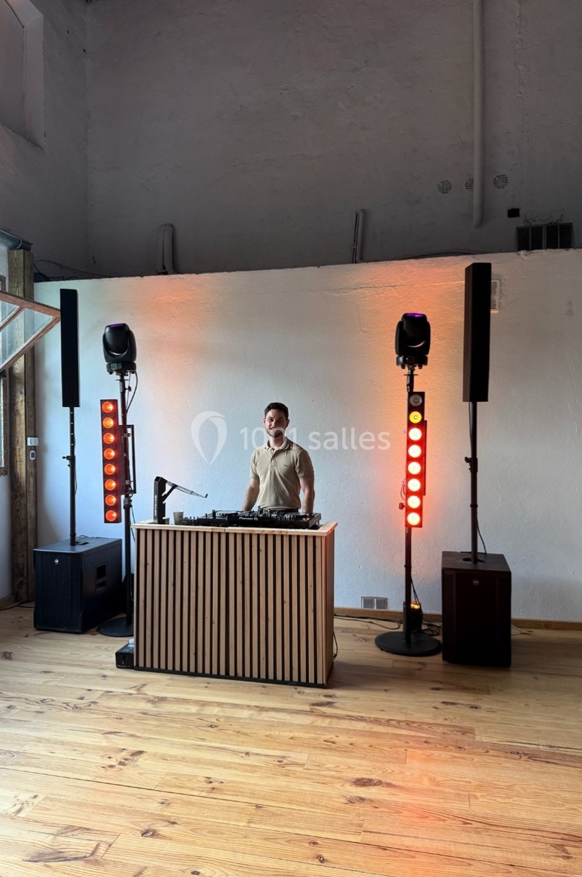 Un DJ joue de la musique derrière une table équipée, entourée de lumières et d'enceintes dans une salle lumineuse.