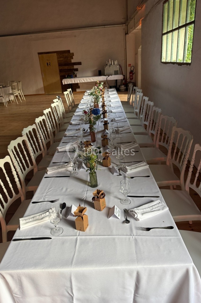 Table longue dressée pour un repas, avec nappes blanches, chaises assorties, décorations florales et petits cadeaux.