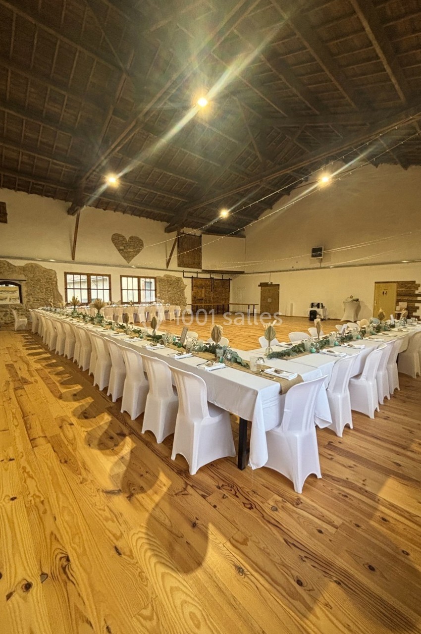 Salle en bois avec de longues tables dressées, nappes blanches, chaises alignées et décoration sobre.