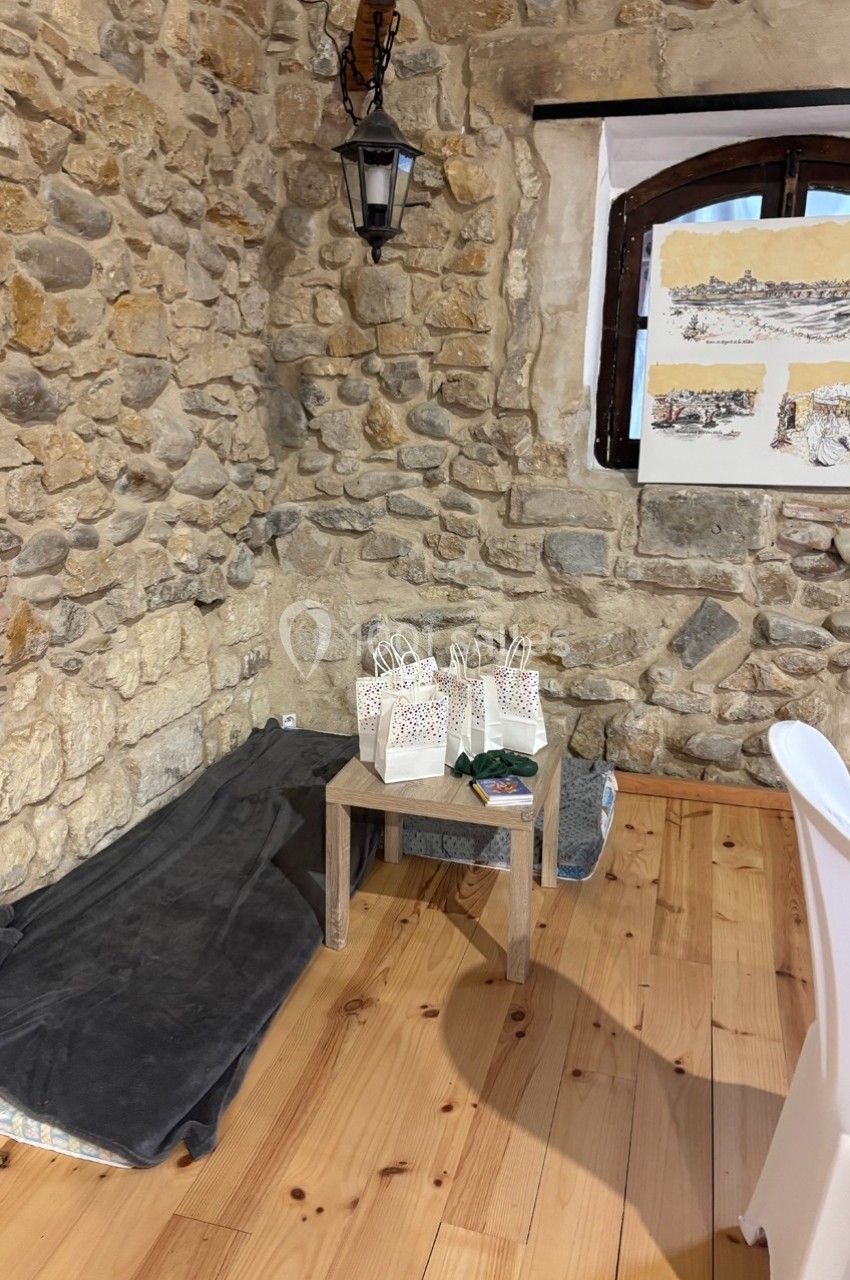 Table en bois avec des sacs cadeaux, tapis et coussins au sol, dans une pièce aux murs en pierre avec une fenêtre et des…