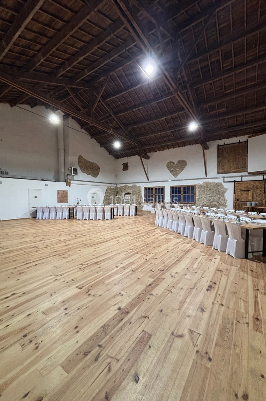 Salle spacieuse avec parquet en bois, tables alignées et chaises blanches, murs en pierre et plafond en poutres apparentes.