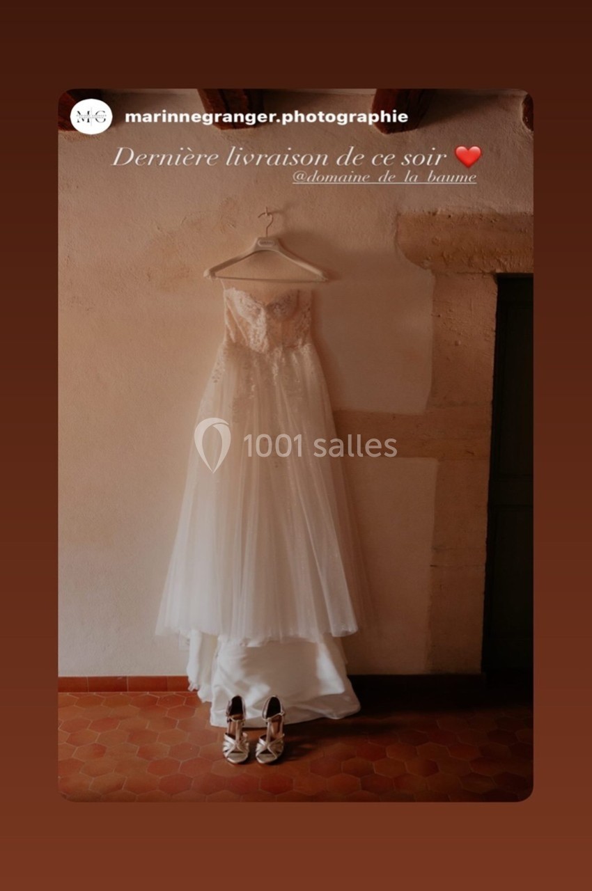 Robe de mariée blanche suspendue à un mur beige, avec des chaussures assorties posées au sol.