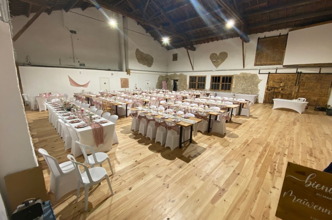 Salle de réception avec tables décorées pour un événement, chaises alignées et plafond en bois apparent.