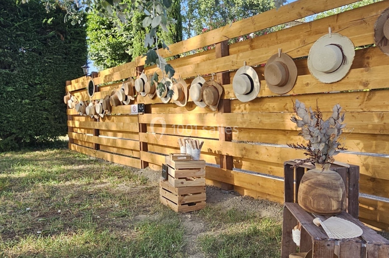 Chapeaux en paille accrochés à une clôture en bois dans un jardin, avec des caisses et des décorations rustiques au sol.