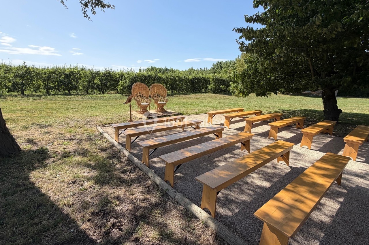 Bancs en bois disposés en rangées en extérieur, face à deux fauteuils en osier sous un arbre, près d'un verger.