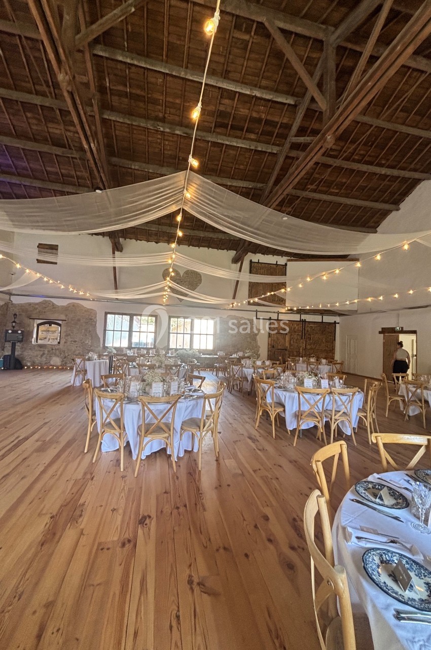 Salle de réception avec tables dressées, chaises en bois, guirlandes lumineuses et plafond en poutres apparentes.