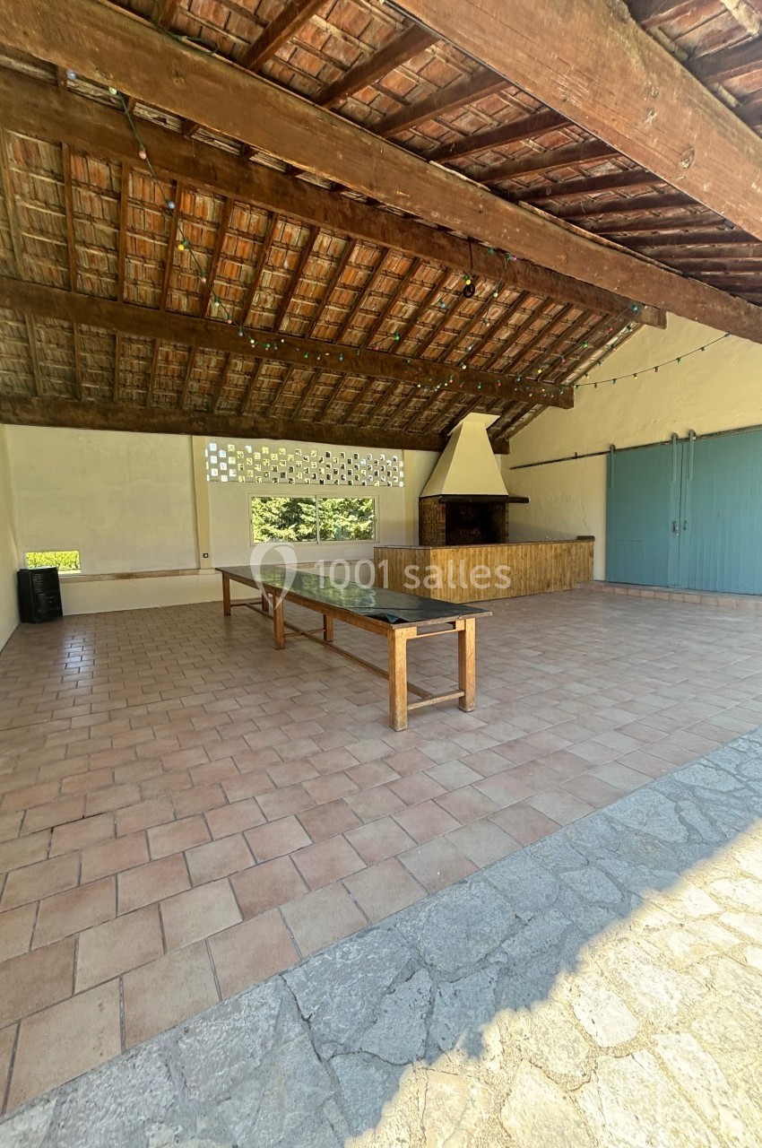 Abri ouvert avec toit en tuiles, grande table en bois, barbecue en pierre et portes bleues sur un sol carrelé.