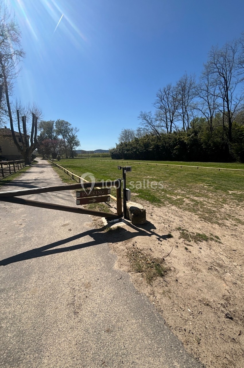 Barrière en bois fermant l'accès à un parking herbeux, entouré d'arbres sous un ciel dégagé.