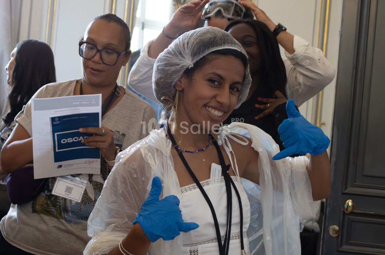 Une jeune femme souriante en robe blanche, coiffée d'une charlotte et de gants bleus en latex, pose avec deux pouces levés devant un groupe de personnes dans une pièce aux boiseries claires. Wait, let me check constraints: no "Photo de", under 125 chars.