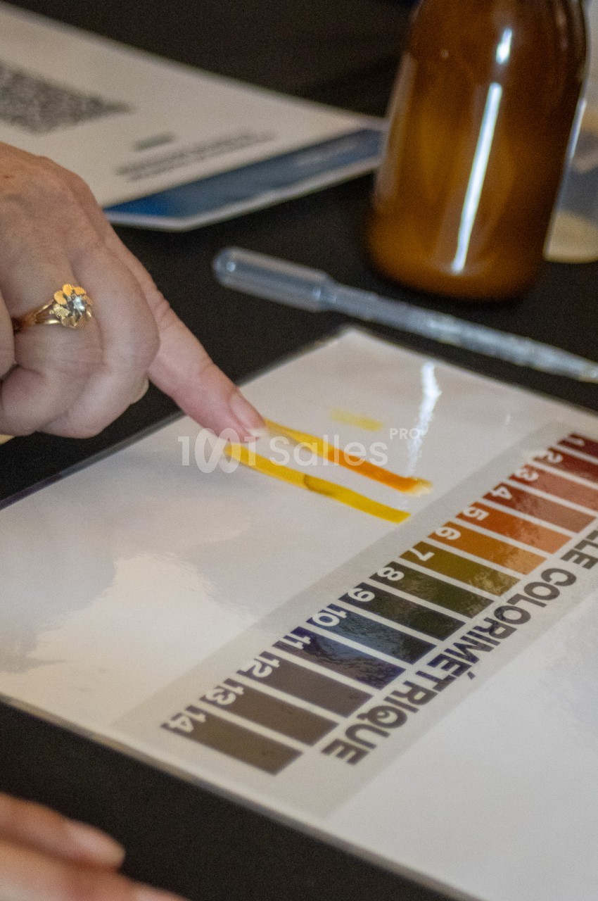 Une main pointe une bande de liquide jaune-orangé déposée sur une feuille blanche, à côté d'une échelle colorimétrique de pH et d'une pipette en plastique.
