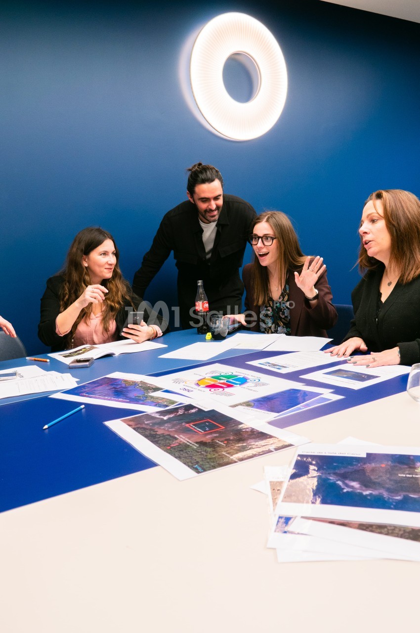 Quatre personnes échangent autour d'une grande table bleue couverte de documents imprimés et de cartes, sous un grand luminaire circulaire sur mur bleu.