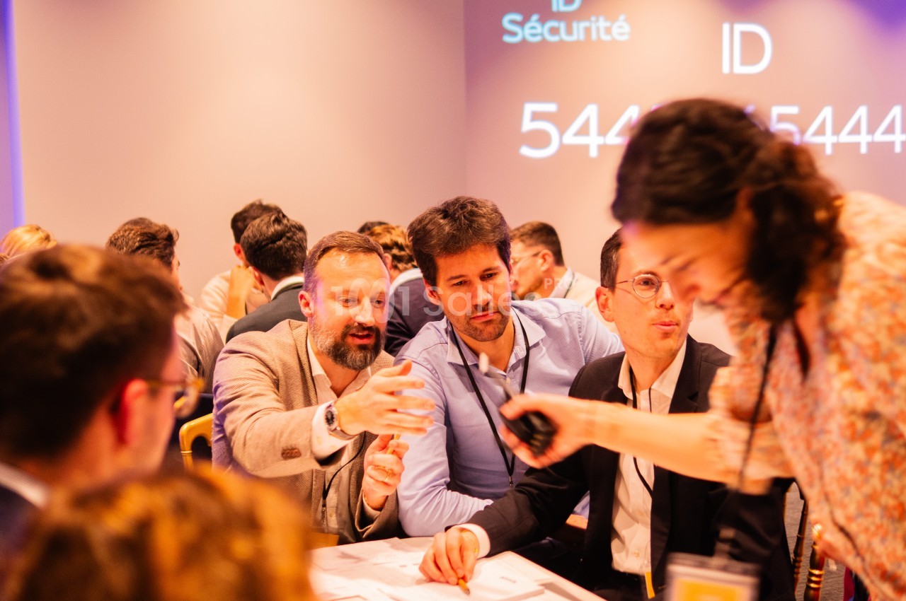 Des participants en tenue professionnelle réunis autour d'une table lors d'un atelier, sous un éclairage chaud, avec un écran en fond affichant « ID Sécurité ». Wait: I must not transcribe text incrusted in the image (enseigne, affiche). The "ID