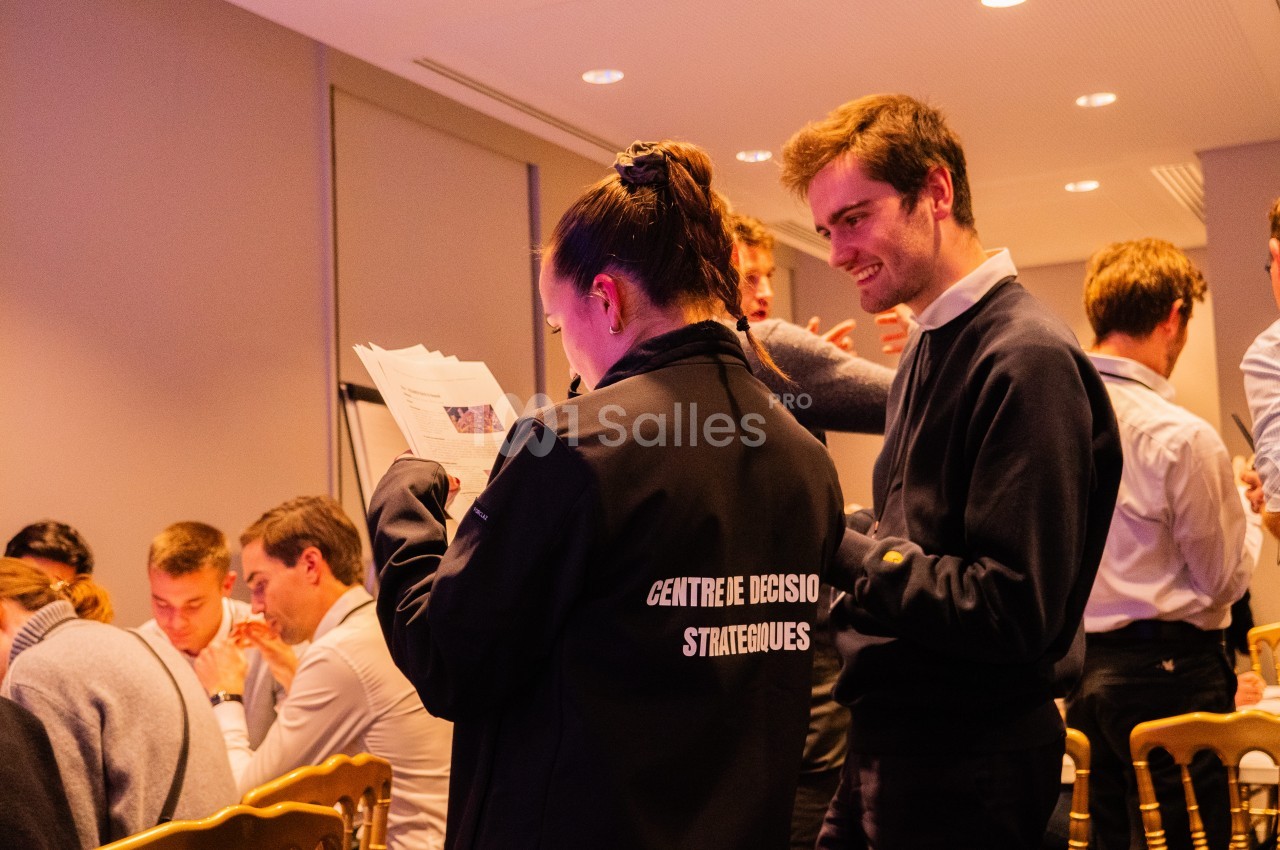 Une jeune femme de dos, en veste noire, lit un document près d'un homme souriant en pull sombre, dans une salle de réception aux chaises dorées où d'autres participants sont assis.