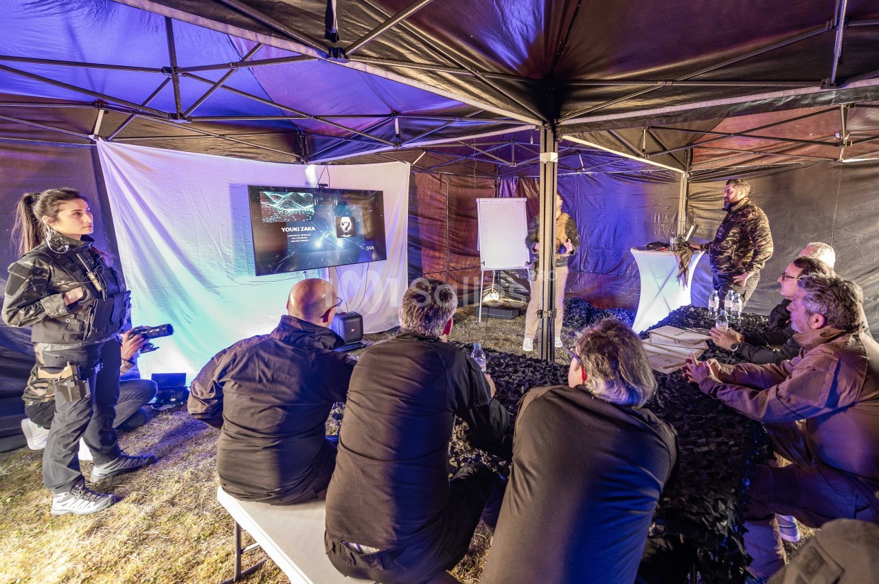 Sous une tente noire éclairée en bleu, un groupe de participants assis écoute un formateur en tenue camouflage devant un écran de projection, tandis qu'une femme en tenue militaire se tient à gauche. Let me check length: roughly 190 characters, too long.