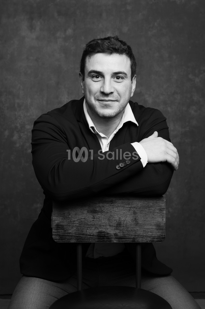 Portrait en noir et blanc d'un homme souriant, assis sur une chaise en bois, les bras croisés sur le dossier, sur fond sombre texturé.