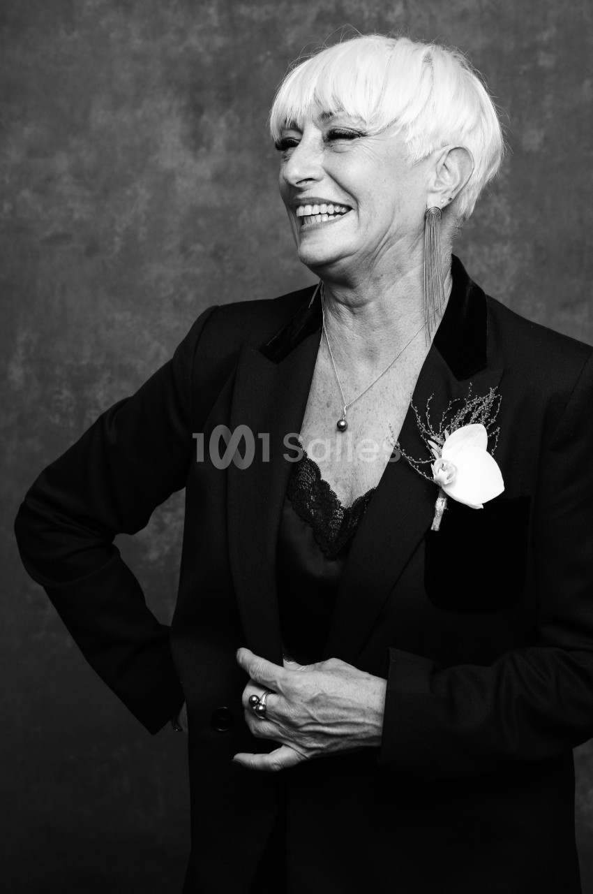 Portrait en noir et blanc d'une femme aux cheveux courts blancs, souriante, en veste de smoking avec une fleur blanche au revers, sur fond gris texturé.