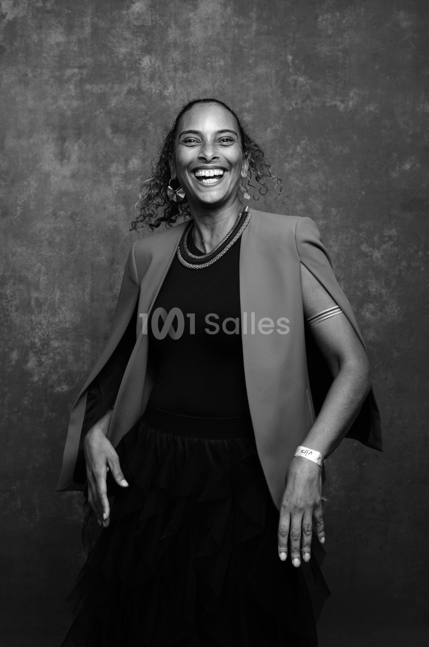 Portrait en noir et blanc d'une femme souriante, debout devant un fond texturé gris, vêtue d'une veste sans manches et d'une jupe noire à volants.